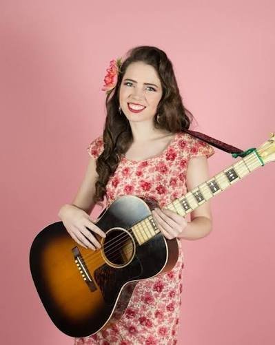 Martha Spencer smiling while holding an acoustic guitar, wearing a floral dress and flower in her hair against a pink background