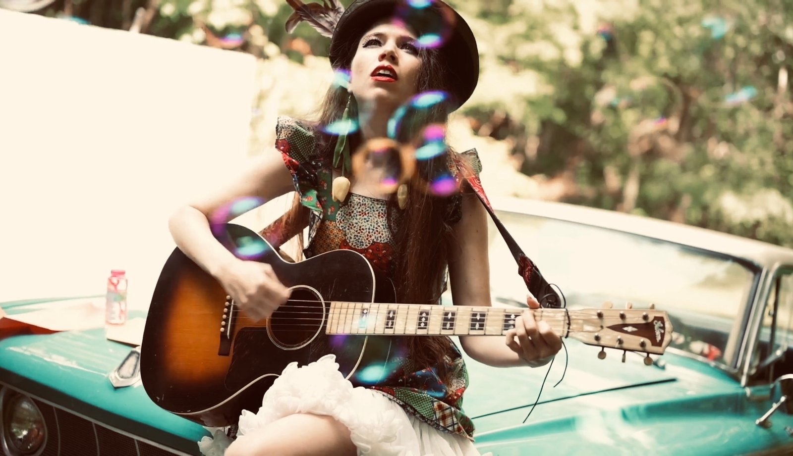 Martha Spencer playing acoustic guitar while seated on a turquoise vintage car, with colorful bokeh light reflections and outdoor trees behind her